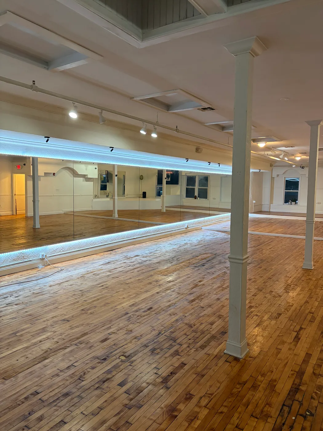 The full length mirrored wall and hardwood floor of the studio, lit by a soft LED strip
