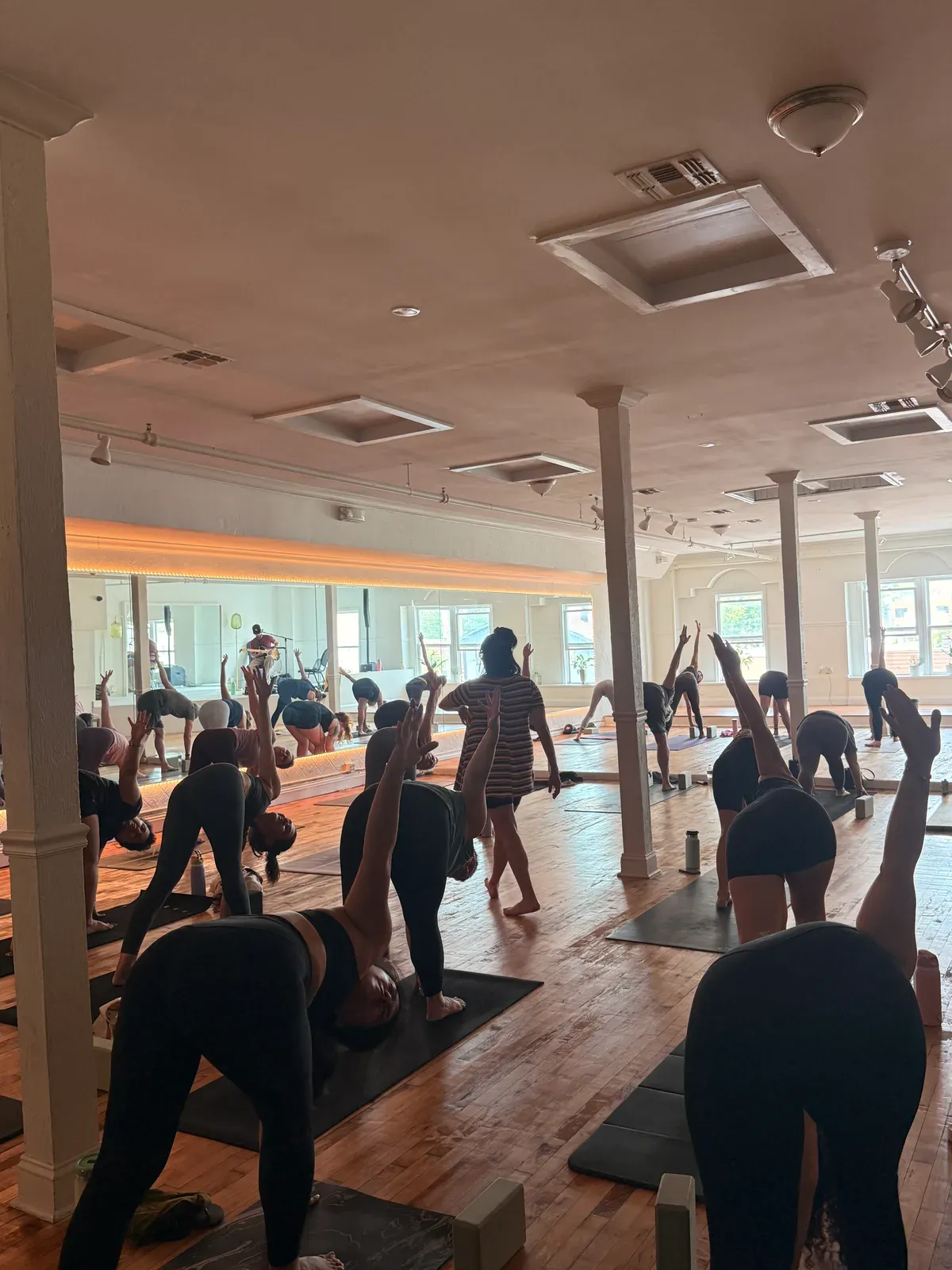 A packed yoga class mid flow on the main floor, arms reaching toward the mirrored wall