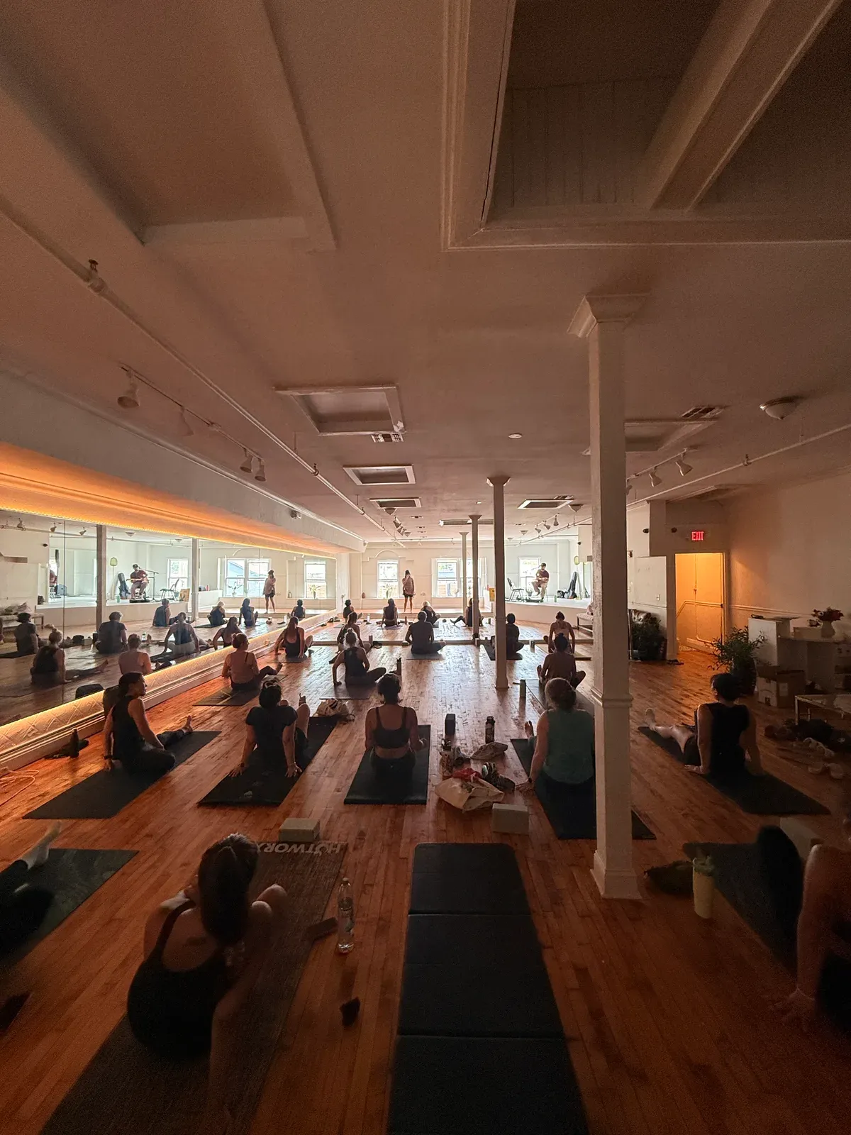 A full class seated on mats across the main floor at golden hour, facing the mirrored wall